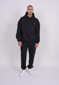 Mustat hupparit ja collegehousut ovat valmistettu puuvillasekoitteesta, ja niissä on etutasku sekä valkoiset tennarit. Minimalistinen muotoilu ilman kuvioita.