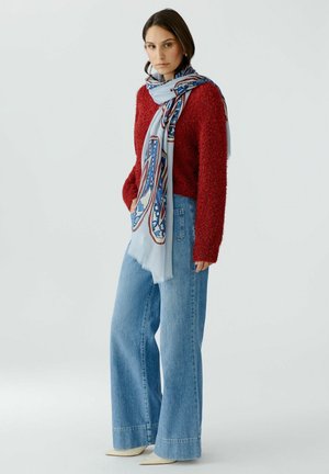 Vrouw die een rode trui met structuur draagt, een lichtblauwe sjaal met patroon, wijde blauwe spijkerbroek en witte puntige schoenen, staand tegen een effen achtergrond.