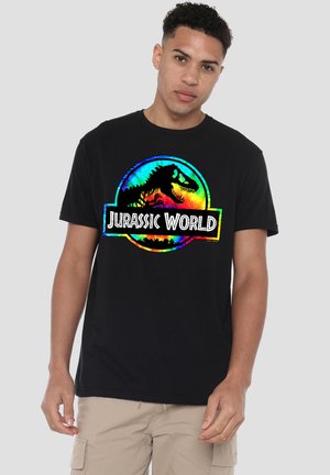 Junger Mann trägt ein schwarzes Jurassic World T-Shirt mit buntem Dinosaurierlogo vorne, steht vor einfarbigem Hintergrund.
