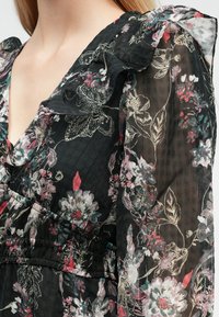 Čierne kvetinové šaty s volánovým výstrihom a rukávmi. Na priesvitnej látke so štruktúrovaným základom výrazné ružové a zelené kvetinové vzory.