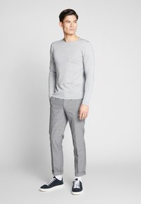 Lys grå langermet skjorte, kombinert med grå rutete bukser og marineblå sneakers. Modellen står med hendene i lommene, foran en nøytral bakgrunn.