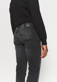 Person wearing dark gray jeans with stitched back pockets and a black long-sleeve shirt, standing against a plain light background.