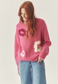 Pulover tricotat roz cu broderie de flori în roșu, crem și alb. Prezentând un croi lejer și tiv ribat. Asortat cu blugi de culoare albastru deschis.