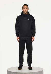Man staat op een rond platform, gekleed in een zwarte hoodie, bijpassende zwarte broek met zakken en zwarte sneakers met gumzolen.