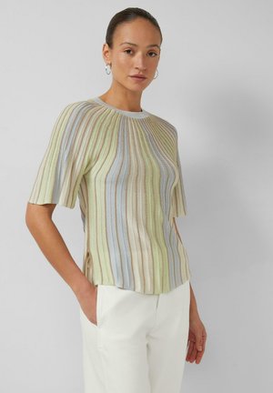 Frau trägt pastellfarben gestreiftes kurzärmeliges Strickoberteil und weiße Hose, steht mit einer Hand in der Tasche vor einem einfarbigen Hintergrund.