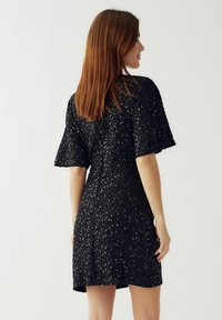 Juoda suknelė, pagaminta iš blizgučių audinio, su trumpomis, platėjančiomis rankovėmis ir prigludusia siluetu, su kelio ilgio krašto ir blizgančiais akcentais.