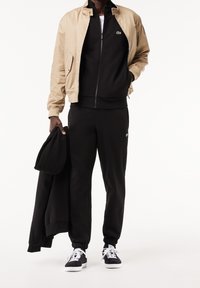 Svart träningsdräkt med jacka med dragkedja, beige bomberjacka och svarta sneakers med vita sulor. Har en logotyp på jackan och byxorna.