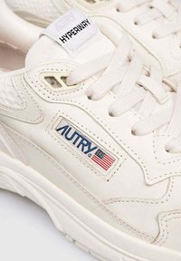 Sneakers in pelle bianca con dettagli in rete, caratterizzati da un prominente logo blu "AUTRY" e un particolare bandiera americana di lato. Lacci piatti e suola testurizzata.