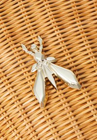 Zlatý kovový hmyzový brož s geometrickými tvary, leštěným povrchem a dvěma velkými křídly, umístěný na tkaném slámovém pozadí.