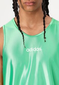 Top sportiv verde, fabricat dintr-un material lucios, cu guler rotund și logo-ul alb "adidas" centrat pe piept.