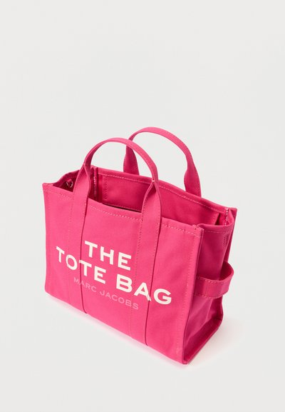 Ροζ καμβάς τσάντα tote με δύο λαβές στο πάνω μέρος και κλείσιμο με κουμπί στο πλάι. Περιέχει μεγάλα λευκά γράμματα που αναγράφουν "Η ΤΣΑΝΤΑ TOTE" και "MARC JACOBS."