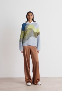 Pull en tricot multicolore avec des motifs en vagues bleues, vertes et jaunes. Assorti avec un pantalon large marron et des baskets blanches.