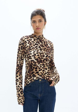 Mujer con cabello oscuro recogido, vistiendo una camiseta de manga larga con estampado de leopardo y vaqueros azul oscuro sobre un fondo blanco liso.