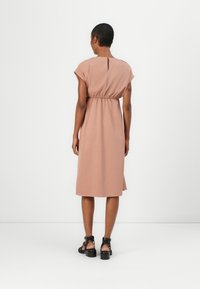 Blush pink dress with short sleeves, cinched waist, and back keyhole opening, falling just below the knee. Worn with black strappy sandals.