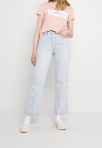 Nainen, joka on pukeutunut vaaleansinisiin korkeavyötäröisiin farkkuihin, vaaleanpunaiseen Levi's-t-paitaan, valkoisiin sukkiin ja beigekirjaviin lenkkarieksiin, seisoo käsi taskussa.