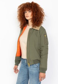 Donna con i capelli rossi ricci indossa una giacca bomber verde oliva con fodera arancione, un top beige e jeans blu, in piedi contro uno sfondo bianco.