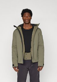 Olivovo zelená nepremokavá bunda s kapucňou, zapínaním na zips, textúrovanou tkaninou a elastickými manžetami. Nosená cez čiernu košeľu s dlhým rukávom a zodpovedajúce nohavice.