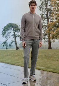 Grey zip-up sweater with ribbed cuffs, paired with light grey jeans and beige sneakers, set against a misty outdoor backdrop with trees.