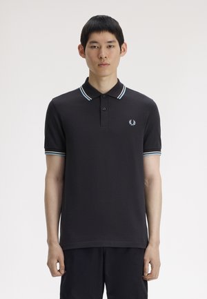 Joven con una camiseta tipo polo negra con cuello y puños de manga a rayas en azul claro y blanco, posando frente a un fondo blanco liso.