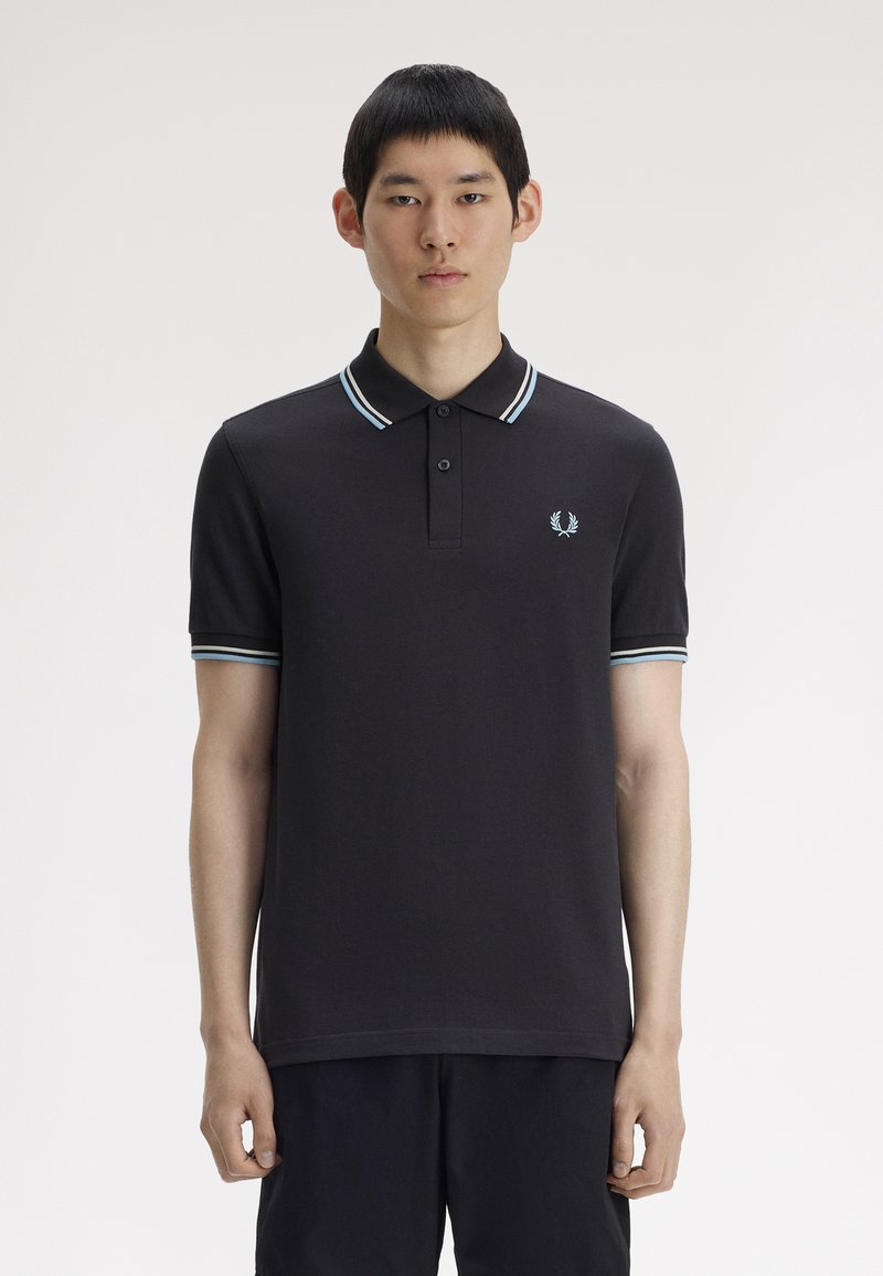 Jeune homme portant un polo noir avec un col et des poignets de manches rayés bleu clair et blanc, debout devant un fond blanc uni.