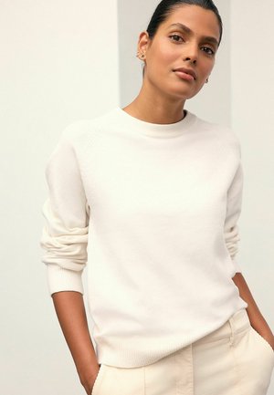 Sweatshirt alb cu guler rotund și manșete cu dungi, având o textură moale. Modelul îl asortează cu pantaloni bej deschis.