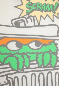 T-shirt met een groene cartoonpersoonlijkheid met oranje accenten, grijze contouren en de tekst "SCRAM!" in vet groen, alles op een lichte achtergrond.