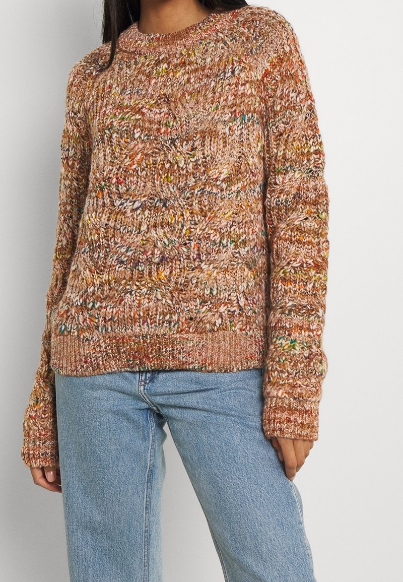 Femme portant un pull en maille épaisse multicolore avec un motif torsadé et un jean denim bleu clair sur un fond uni.