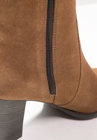 Bottine en daim marron avec fermeture éclair latérale noire et talon bloc en bois empilé sur fond clair.