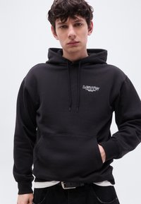 Νεαρός άνδρας που φοράει μαύρο φούτερ Levi’s με το λογότυπο στο στήθος, με το ένα χέρι στην τσέπη, στέκεται μπροστά σε απλό ανοιχτόχρωμο φόντο.