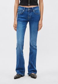 Kék farmernadrág bővülő szárakkal, kifakult textúrával. Öt zsebbel és elöl gombos rögzítéssel rendelkezik. Fekete, hegyes orrú cipővel párosítva.