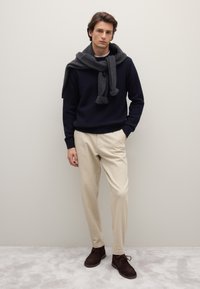 Maglione lavorato a maglia blu navy con sciarpa grigia avvolta intorno al collo, abbinato a pantaloni beige e stivaletti in camoscio marrone, su sfondo neutro.