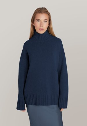Woman with long blond hair wearing a dark blue ribbed turtleneck sweater and a blue skirt, facing forward against a plain background.