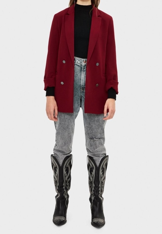 Personne portant un blazer croisé rouge foncé, un haut noir, un jean gris délavé à l'acide et des bottes de cowboy noires brodées, debout devant un fond uni.