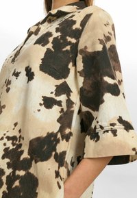 Beige shirt met zwart en bruin koeienprintdesign, met een kraag en korte, brede mouwen. Zachte textuur, met een ontspannen pasvorm.