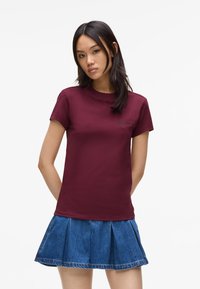 Tricou din bumbac burgundy, cu mâneci scurte, decolteu rotund și un logo discret în negru. Asortat cu o fustă plisată din denim.