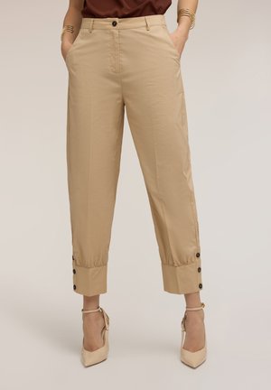 Pantalon court beige taille haute avec poignets boutonnés, porté avec des talons beiges à bride cheville, mains dans les poches et bracelets dorés aux poignets.