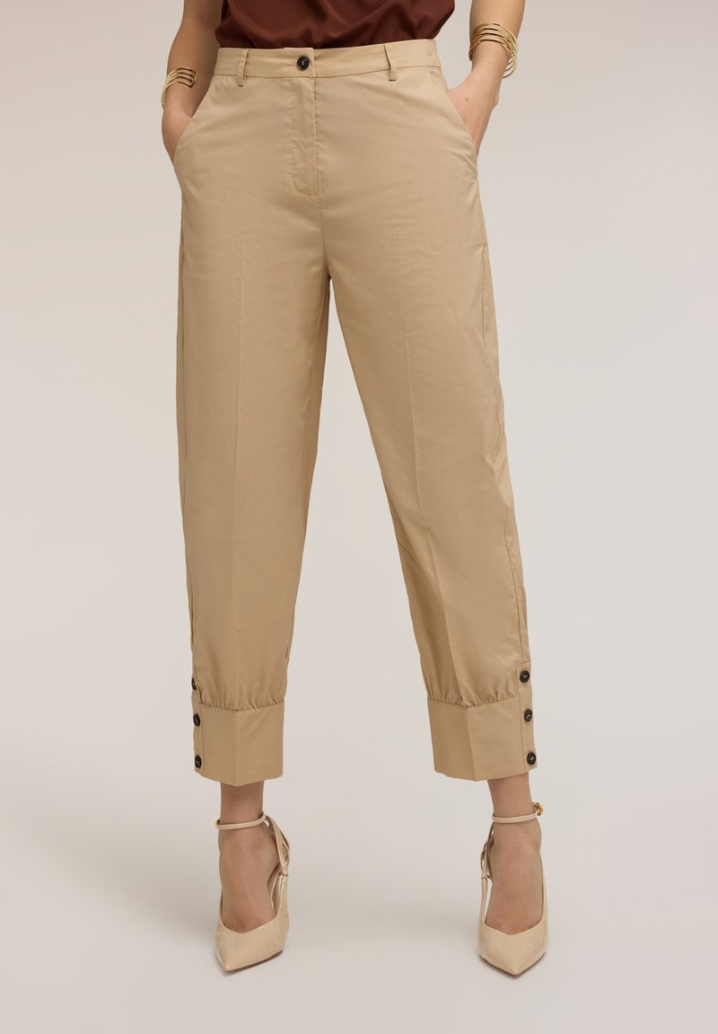 Pantalones cortos beige de cintura alta con puños con botones, usados con zapatos de tacón beige con tira en el tobillo, manos en los bolsillos y pulseras doradas en las muñecas.