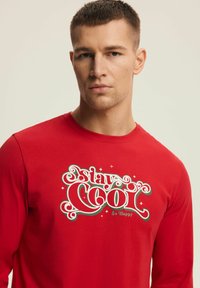 Czerwona koszulka z długim rękawem z białym i zielonym wzorem graficznym oraz napisem „Stay Cool & Be Happy” falistą czcionką.