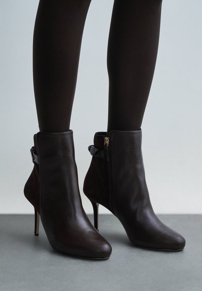 Botines de cuero marrón con punta puntiaguda, tacón de aguja delgado, cremallera lateral y una correa decorativa en el tobillo.