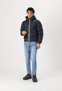 Giacca puffer blu navy con cappuccio, caratterizzata da una zip gialla. Indossata con jeans azzurri e sneakers nere. Texture liscia e design a blocchi.