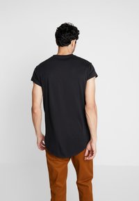 Black short-sleeve t-shirt with a rounded hem, crafted from soft fabric; worn with brown pants, showcasing a simple, minimalist design.