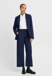 Dunkelblauer Anzug mit weit geschnittenen Hosen und einem Ein-Knopf-Blazer. Getragen über einem weißen Hemd mit Kragen, kombiniert mit schwarzen Slippern.