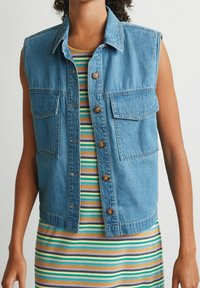 Gilet sans manches en denim bleu avec deux poches avant et des boutons en cuivre, superposé à une robe côtelée à rayures multicolores dans les tons vert, jaune et violet.