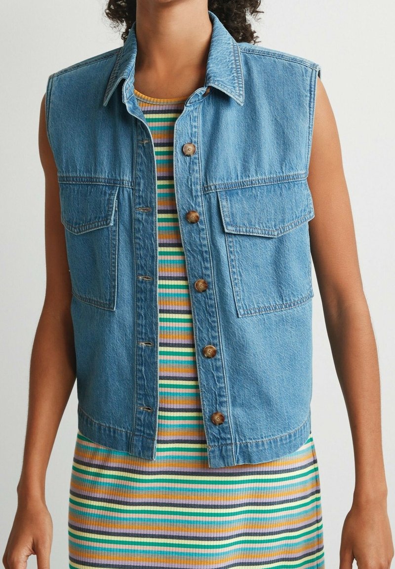 Gilet sans manches en denim bleu avec deux poches avant et des boutons en cuivre, superposé à une robe côtelée à rayures multicolores dans les tons vert, jaune et violet.