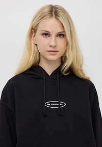 Czarna bluza z kapturem posiadająca przednią kieszeń i sznurki. Na środku białego tekstu znajduje się napis "BĄDŹ ORYGINALNY" w kształcie owalu. Miękka faktura materiału.