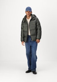 Zelena puffer jakna s kapuljačom, bež polo majica, plave traperice i crne cipele. Model nosi plavu kapu, u stojećem položaju.