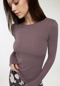 Długi rękaw, prążkowany top w kolorze mauve, z okrągłym dekoltem i dopasowanym krojem. Model trzyma dolną część, prezentując gładką fakturę.