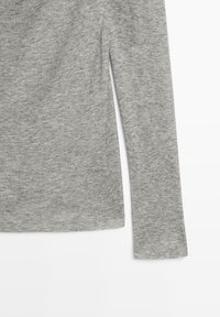 Détail d'une manche et d'une couture latérale d'un pull en maille gris chiné à manches longues, tissu doux et texturé sur un fond blanc.