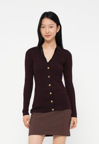 Young woman with straight black hair wearing a dark purple ribbed cardigan with gold buttons and a brown skirt, standing against a white background.
