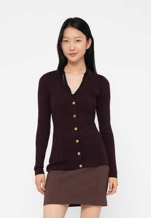 Young woman with straight black hair wearing a dark purple ribbed cardigan with gold buttons and a brown skirt, standing against a white background.